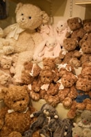 A collection of plush teddy bears and stuffed animals are arranged on shelves. The assortment includes brown and beige teddy bears of various sizes, some wearing bows. There are also pink and grey stuffed animals among the collection.