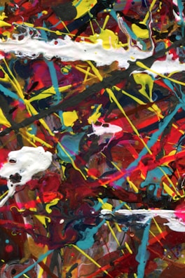 A vibrant composition of swirling colors including red, yellow, blue, and white, with splashes and drips creating a chaotic abstract appearance. Thick, expressive strokes and overlapping layers of paint form an energetic visual texture.