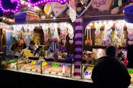 A brightly lit candy and snack stall at a fair or market features an array of colorful treats. Heart-shaped gingerbread cookies with messages hang prominently above the stall, while various nuts, popcorn, and sweets are displayed in containers. Several people are either working behind the counter or browsing, one customer facing away from the camera.