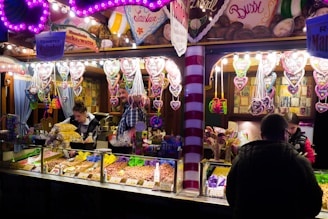 A brightly lit candy and snack stall at a fair or market features an array of colorful treats. Heart-shaped gingerbread cookies with messages hang prominently above the stall, while various nuts, popcorn, and sweets are displayed in containers. Several people are either working behind the counter or browsing, one customer facing away from the camera.