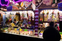 A brightly lit candy and snack stall at a fair or market features an array of colorful treats. Heart-shaped gingerbread cookies with messages hang prominently above the stall, while various nuts, popcorn, and sweets are displayed in containers. Several people are either working behind the counter or browsing, one customer facing away from the camera.