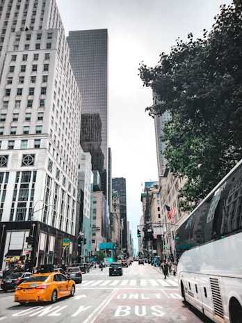 A busy city street lined with tall buildings and bustling with traffic. A yellow taxi and several cars are visible on the road, along with a large bus. Pedestrians are walking on sidewalks, and the scene captures a mix of urban life with elements such as traffic signals, street signage, and greenery from trees.