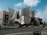 A modern, metallic building with an intricate and unconventional design dominates the scene. The structure has smooth, curved surfaces and sharp angles. In the foreground, there is a street with cars and pedestrians crossing. Adjacent to the building is a large advertisement featuring a person associated with LA Phil. The sky is partly cloudy with patches of blue showing through.