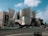 A modern, metallic building with an intricate and unconventional design dominates the scene. The structure has smooth, curved surfaces and sharp angles. In the foreground, there is a street with cars and pedestrians crossing. Adjacent to the building is a large advertisement featuring a person associated with LA Phil. The sky is partly cloudy with patches of blue showing through.