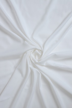A delicate, smooth white fabric is elegantly draped and gathered in a swirling pattern, creating soft folds and flowing lines across the surface.