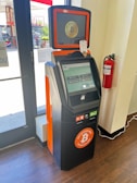 A Bitcoin ATM with an orange and black design is located indoors on a wooden floor. The machine has a touchscreen display and is positioned next to a large window and fire extinguisher. Visible on the screen is a buy and sell Bitcoin interface. The ATM has signage promoting Bitcoin transactions.