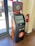 A Bitcoin ATM with an orange and black design is located indoors on a wooden floor. The machine has a touchscreen display and is positioned next to a large window and fire extinguisher. Visible on the screen is a buy and sell Bitcoin interface. The ATM has signage promoting Bitcoin transactions.