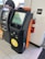 A Bitcoin ATM is situated next to a traditional ATM in a store or public area. The Bitcoin ATM has a screen displaying an interface for digital transactions, with a prominent logo indicating Bitcoin usage. An 'EXIT' sign is on the wall nearby, alongside a fire extinguisher mounted on the wall.