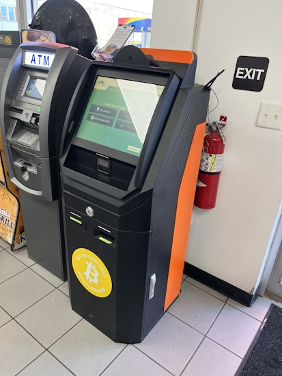 A Bitcoin ATM is situated next to a traditional ATM in a store or public area. The Bitcoin ATM has a screen displaying an interface for digital transactions, with a prominent logo indicating Bitcoin usage. An 'EXIT' sign is on the wall nearby, alongside a fire extinguisher mounted on the wall.