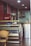 The image depicts the interior of a small eatery or café. There is a refrigerated display case with some items inside and on top. A wicker chair is placed in front of the display case. To the left, there's a Coca-Cola refrigerator. The back of the room features a stainless steel kitchen hood and some kitchen equipment.