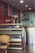 The image depicts the interior of a small eatery or café. There is a refrigerated display case with some items inside and on top. A wicker chair is placed in front of the display case. To the left, there's a Coca-Cola refrigerator. The back of the room features a stainless steel kitchen hood and some kitchen equipment.
