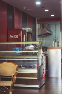 The image depicts the interior of a small eatery or caf&eacute;. There is a refrigerated display case with some items inside and on top. A wicker chair is placed in front of the display case. To the left, there's a Coca-Cola refrigerator. The back of the room features a stainless steel kitchen hood and some kitchen equipment.