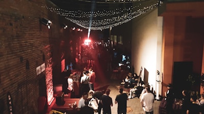 An alleyway filled with people appears to be hosting an event or gathering. Strings of twinkling lights are hanging overhead, creating a festive atmosphere. The scene is illuminated with warm colors, with red and orange lighting reflecting off the brick wall on the left. A group of people are gathered around various seating arrangements, engaging in conversation and mingling. There's a bright spotlight in the distance, possibly indicating a stage or focal point of the event.