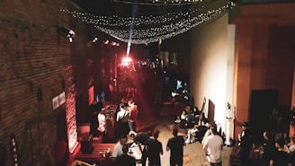 An alleyway filled with people appears to be hosting an event or gathering. Strings of twinkling lights are hanging overhead, creating a festive atmosphere. The scene is illuminated with warm colors, with red and orange lighting reflecting off the brick wall on the left. A group of people are gathered around various seating arrangements, engaging in conversation and mingling. There's a bright spotlight in the distance, possibly indicating a stage or focal point of the event.