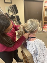 A person with dark hair wearing a red sweater is examining the back and neck of another person with blonde hair in a ponytail. The person being examined is wearing a medical gown and sitting on a medical chair. The background shows medical equipment and certificates on the wall.