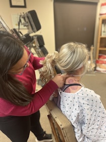 A person with dark hair wearing a red sweater is examining the back and neck of another person with blonde hair in a ponytail. The person being examined is wearing a medical gown and sitting on a medical chair. The background shows medical equipment and certificates on the wall.