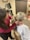 A person with dark hair wearing a red sweater is examining the back and neck of another person with blonde hair in a ponytail. The person being examined is wearing a medical gown and sitting on a medical chair. The background shows medical equipment and certificates on the wall.