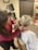 A person with dark hair wearing a red sweater is examining the back and neck of another person with blonde hair in a ponytail. The person being examined is wearing a medical gown and sitting on a medical chair. The background shows medical equipment and certificates on the wall.