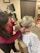 A person with dark hair wearing a red sweater is examining the back and neck of another person with blonde hair in a ponytail. The person being examined is wearing a medical gown and sitting on a medical chair. The background shows medical equipment and certificates on the wall.