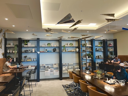 A modern lounge area features a long wooden table surrounded by chairs, with a few people working on laptops. The back wall showcases tall bookshelves decorated with books, plants, and various decorative items. Paper airplanes hang from the ceiling, adding a playful element. The space is well-lit with both natural and artificial lighting.