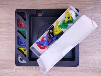 A wooden surface holding a black tray and a partially opened white box containing colorful plastic corner pieces in red, blue, green, and yellow. The tray also has slots with these same colored pieces neatly organized.