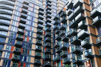 A modern high-rise residential building with numerous glass balconies, showcasing an intricate pattern of alternating windows and colorful panels. The architecture is striking and geometric, with a combination of blue, brown, and orange elements, creating a dynamic facade.