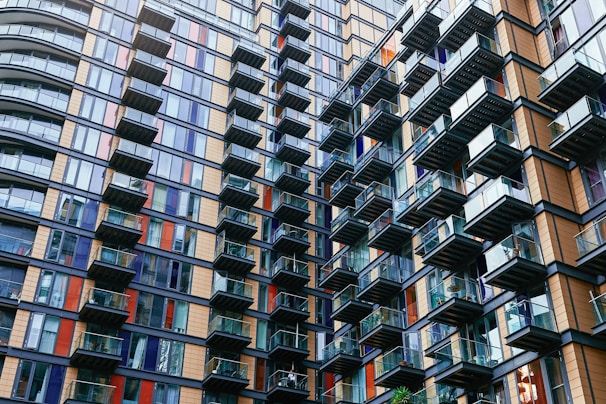 A modern high-rise residential building with numerous glass balconies, showcasing an intricate pattern of alternating windows and colorful panels. The architecture is striking and geometric, with a combination of blue, brown, and orange elements, creating a dynamic facade.