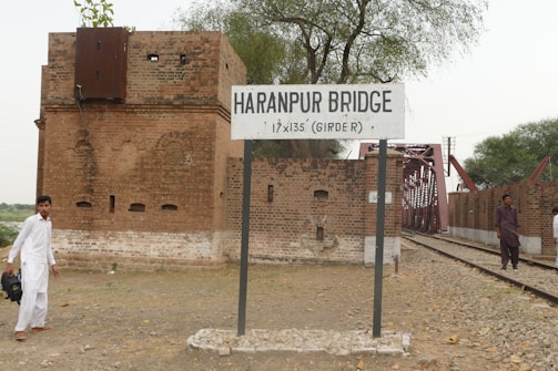 A railway bridge sign reads 'Haranpur Bridge' in front of a red brick building. Train tracks run alongside the building and into the distance. Two individuals are walking beside the tracks, one in white traditional attire and the other in darker clothing. The area is surrounded by sparse greenery and trees.