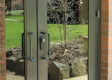 Glass double doors with black handles reflect a view of a garden outside. The garden contains a small tree with bare branches and a few blooms, surrounded by rocks and mulch. Bright green grass and a path are visible in the background. A brick wall frames one side of the doors.