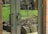 Glass double doors with black handles reflect a view of a garden outside. The garden contains a small tree with bare branches and a few blooms, surrounded by rocks and mulch. Bright green grass and a path are visible in the background. A brick wall frames one side of the doors.