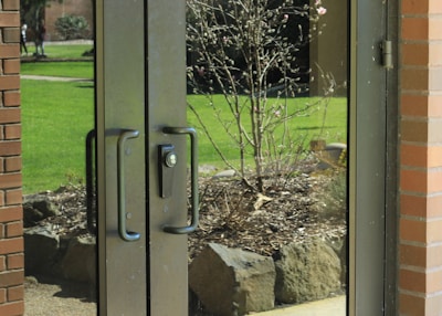 Glass double doors with black handles reflect a view of a garden outside. The garden contains a small tree with bare branches and a few blooms, surrounded by rocks and mulch. Bright green grass and a path are visible in the background. A brick wall frames one side of the doors.