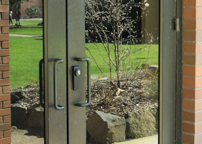 Glass double doors with black handles reflect a view of a garden outside. The garden contains a small tree with bare branches and a few blooms, surrounded by rocks and mulch. Bright green grass and a path are visible in the background. A brick wall frames one side of the doors.