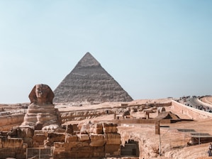 A large pyramid dominates the background, with the iconic Sphinx in the foreground. The setting is an ancient desert landscape with stone ruins and pathways leading tourists through the site.
