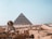 A large pyramid dominates the background, with the iconic Sphinx in the foreground. The setting is an ancient desert landscape with stone ruins and pathways leading tourists through the site.
