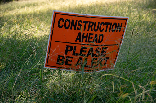 A bright orange warning sign with black lettering is partially obscured by tall grass. The sign reads 'Construction Ahead, Please Be Alert.' The surroundings appear to be an overgrown grassy area.