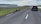 A rural road with several sheep walking along it, while a black car is parked on the opposite side. The landscape is expansive, featuring grassy plains and distant cliffs under a cloudy sky.