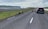 A rural road with several sheep walking along it, while a black car is parked on the opposite side. The landscape is expansive, featuring grassy plains and distant cliffs under a cloudy sky.