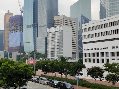The image features a cityscape with multiple modern skyscrapers. Prominently showcased is a white building with 'GENERAL POST OFFICE' written on the front. Nearby, there are high-rise buildings with glass exteriors and a construction site with cranes visible. The foreground is lined with green trees and parked cars along the street.