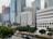 The image features a cityscape with multiple modern skyscrapers. Prominently showcased is a white building with 'GENERAL POST OFFICE' written on the front. Nearby, there are high-rise buildings with glass exteriors and a construction site with cranes visible. The foreground is lined with green trees and parked cars along the street.