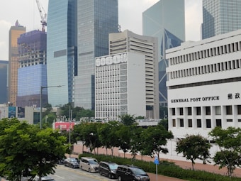 The image features a cityscape with multiple modern skyscrapers. Prominently showcased is a white building with 'GENERAL POST OFFICE' written on the front. Nearby, there are high-rise buildings with glass exteriors and a construction site with cranes visible. The foreground is lined with green trees and parked cars along the street.
