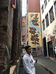 A narrow city alleyway features vibrant street art on a tall building wall, depicting red and white flowers with the word 'Melbourne' on a blue ribbon. A person in a white hoodie stands in the foreground making a hand gesture. The area is surrounded by brick and graffiti-covered buildings.