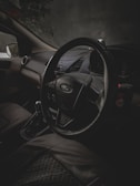 A dimly lit interior of a car focuses on the steering wheel with a Ford logo, surrounded by leather seats and a visible gear shift. The dark, shadowy ambiance suggests an evening or nighttime setting.