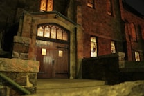 An old stone building is illuminated by warm, ambient lighting, suggesting a historical or possibly religious setting. Stained glass windows with intricate designs are visible, and a large wooden door with iron embellishments is prominently featured.