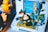 A handheld game console displays a video game scene, surrounded by a creative arrangement of small plants and a figurine resembling a Snorlax character from Pokémon. A bright, yellow rubber duck figure and other Pokémon toys are in the background. The display features pixelated graphics typical of retro video games.