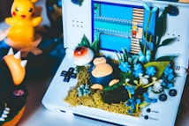 A handheld game console displays a video game scene, surrounded by a creative arrangement of small plants and a figurine resembling a Snorlax character from Pok&eacute;mon. A bright, yellow rubber duck figure and other Pok&eacute;mon toys are in the background. The display features pixelated graphics typical of retro video games.