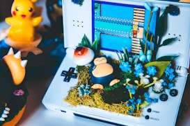 A handheld game console displays a video game scene, surrounded by a creative arrangement of small plants and a figurine resembling a Snorlax character from Pokémon. A bright, yellow rubber duck figure and other Pokémon toys are in the background. The display features pixelated graphics typical of retro video games.