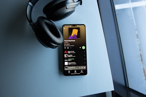 A smartphone displaying a music playlist titled 'Depressing Songs' rests on a light blue surface. Beside it, there is a pair of over-ear headphones. The screen shows a playlist consisting of tracks by various artists, and the display brightness highlights the screen against the muted tones of the setting.