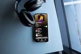 A smartphone displaying a music playlist titled 'Depressing Songs' rests on a light blue surface. Beside it, there is a pair of over-ear headphones. The screen shows a playlist consisting of tracks by various artists, and the display brightness highlights the screen against the muted tones of the setting.