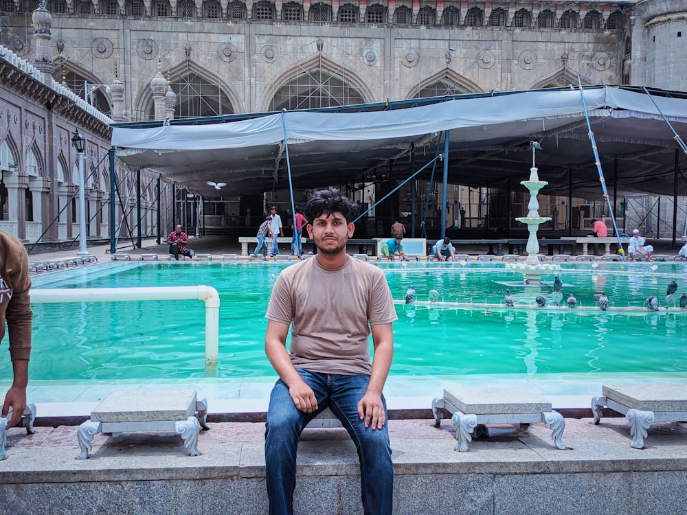 a man sitting on a ledge by a pool with people in the background