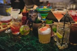 A variety of colorful cocktails arranged on a bar counter, each in a distinct glass type. There are garnishes like fruit slices, mint leaves, and decorative flowers. The setting appears to be a lively bar environment with a mix of classic and tiki-themed drinkware, including a skull-shaped mug.